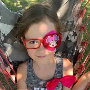Pode incluir: Uma menina pequena usando um tapa-olho vermelho e rosa com um desenho de Minnie Mouse. Ela está usando óculos vermelhos e uma camiseta cinza sem mangas. Ela está sentada em uma rede.