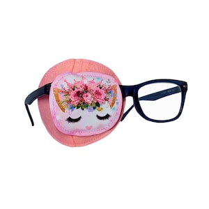 May include: Pink eye patch with a white cat face with a crown and pink flowers. The eye patch is attached to a pair of blue framed glasses.