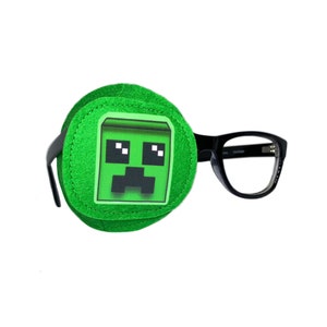 Puede incluir: Un parche para el ojo verde con una cara de Creeper de Minecraft. El parche para el ojo está unido a unas gafas con montura negra.