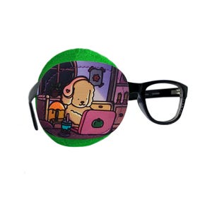 May include: Green eye patch with a cartoon illustration of a dog wearing headphones and using a laptop. The dog is sitting in a room with a window and a bookshelf. The eye patch is attached to a black frame.