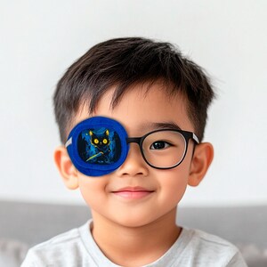 May include: A young child wearing glasses with a blue eye patch featuring a black cat with yellow eyes holding a sword. The eye patch is made of soft fabric and has a white elastic strap.
