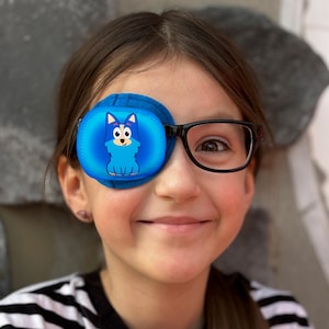 Puede incluir: Una persona joven que lleva gafas y un parche para el ojo azul con un diseño de perro de dibujos animados. El parche para el ojo está asegurado con una correa que va alrededor de la cabeza.