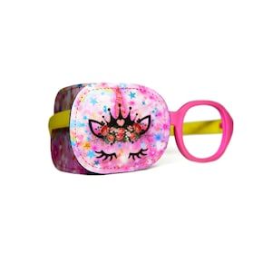 May include: A pink and yellow eye patch with a unicorn design. The unicorn has a crown, flowers, and eyelashes. The patch is made of soft fabric and is designed to be comfortable for children.