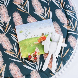 May include: A blue and white play mat with a white pom-pom trim. A book titled "Blowin' in the Wind" by Bob Dylan is on the mat, along with a white stuffed bunny and a wooden toy.