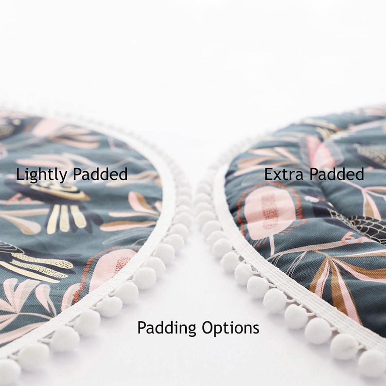 May include: Two round play mats with white pom-pom trim. The mat on the left is labeled "Lightly Padded" and the mat on the right is labeled "Extra Padded". The mats have a green background with a floral pattern and are shown against a white background. Padding Options.