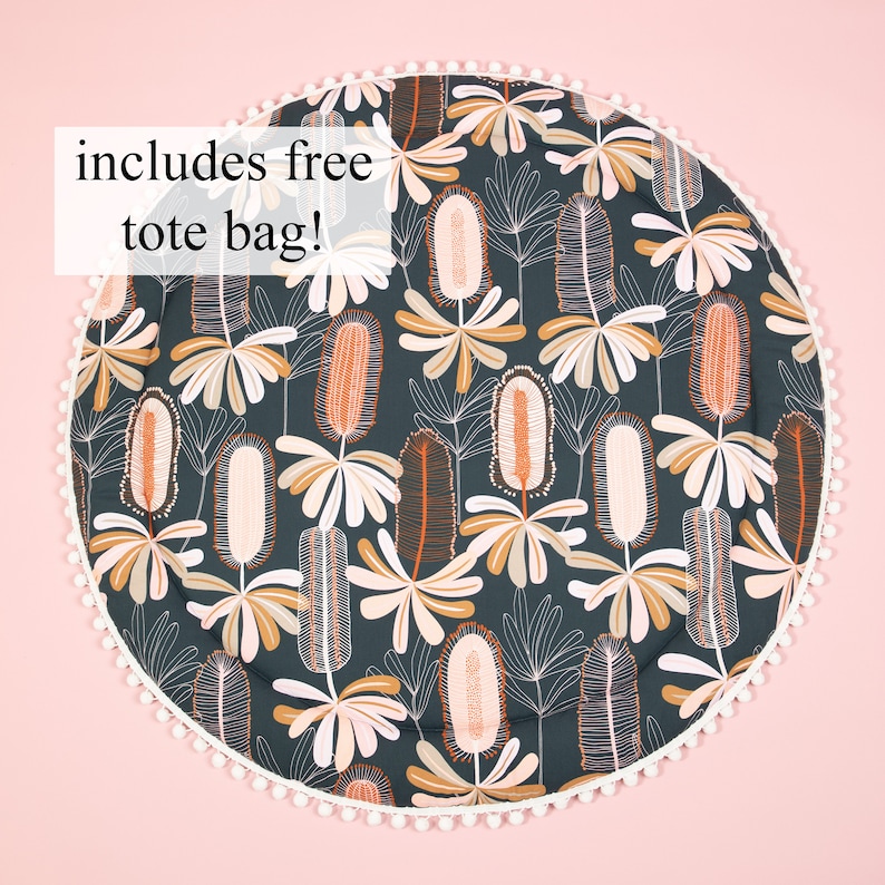 May include: A round play mat with a dark green background and a white pom-pom trim. The mat features a repeating pattern of stylized flowers in shades of orange, pink, and white. The text "includes free tote bag!" is printed in white on a black rectangle at the top of the image.