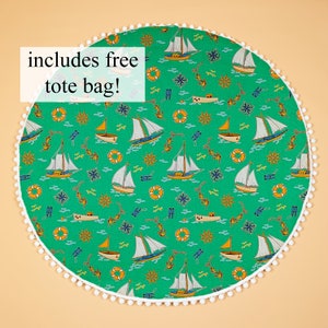 Pode incluir: Um tapete de jogo redondo verde e branco com uma borda de pompons brancos. O tapete de jogo apresenta um padrão repetitivo de veleiros, âncoras e boias salva-vidas. O texto "includes free tote bag!" está impresso no tapete de jogo.