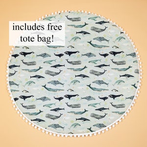 May include: A round, light blue play mat with a white pom-pom trim. The mat features a repeating pattern of whales and other sea creatures. The text "includes free tote bag!" is printed on the mat.