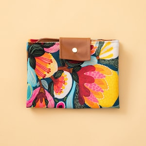 May include: A reusable shopping bag with a floral print featuring bright colours like red, yellow, pink, and blue. The bag has a small brown leather pocket with a gold button.