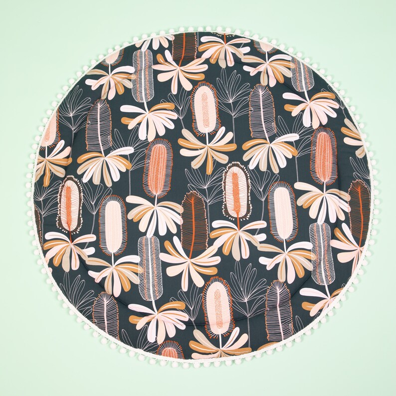May include: A round, dark green play mat with a white pom-pom trim. The mat features a repeating pattern of stylized orange and white flowers.