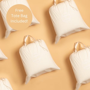 May include: Five white cotton tote bags with a small white logo on each bag. The bags are arranged in a pattern on a light brown background. The text "Free Tote Bag Included!" is written in a white circle on the top left of the image.