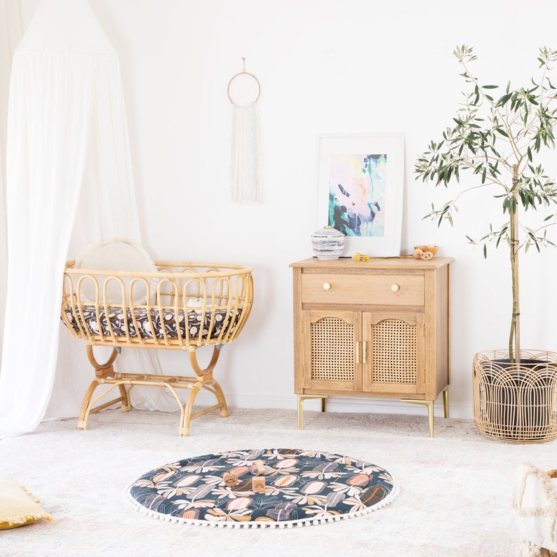 May include: A white canopy hangs over a wicker bassinet with a black and white patterned sheet. A wooden dresser with a woven cane door sits next to the bassinet. A potted olive tree and a round play mat with a floral pattern are on the floor.