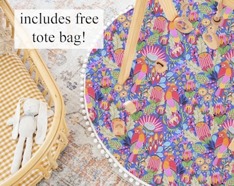 May include: A colorful play mat with a white pom-pom trim and a floral pattern featuring birds and flowers. The mat is on a rug with a wooden play gym in the background. The text "includes free tote bag!" is visible in the image.