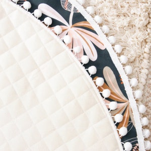 May include: A round, cream-colored play mat with a quilted texture. The mat has a dark green and pink floral print border with a white pom-pom trim.