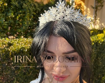 May include: A bride wears a sparkling crystal tiara with a delicate design. The tiara is made of clear crystals and has a pointed crown. The bride is wearing a white dress and has dark hair.