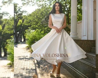May include: A white sleeveless satin dress with a boat neck and a full skirt. The dress is being worn by a model standing on a set of steps. The dress has a small train and the model is wearing white heels.