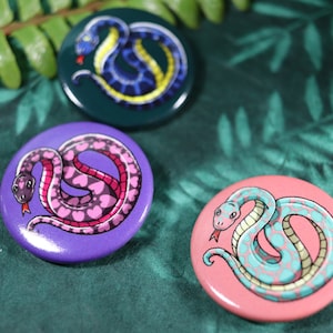 Könnte beinhalten: Drei runde Anstecknadeln mit verschiedenen farbigen Schlangen mit einzigartigen Mustern. Die erste Nadel ist grün mit einer blauen und gelben Schlange. Die zweite Nadel ist lila mit einer pinken und weißen Schlange. Die dritte Nadel ist pink mit einer blauen und weißen Schlange.