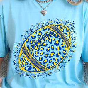 May include: A light blue short-sleeve t-shirt featuring a central graphic of an American football. The football has a yellow and blue leopard print pattern with a distressed texture and is surrounded by a spray of blue and yellow paint splatters.