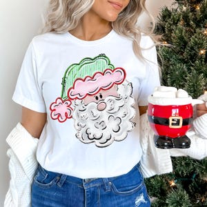 May include: White t-shirt featuring a cartoon Santa Claus face with a pink hat, green trim, and a white beard. A red and black Santa mug filled with marshmallows is held in the hand.