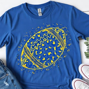May include: A royal blue short-sleeve t-shirt featuring a central graphic of an American football. The football has a yellow and blue leopard print pattern with yellow paint splatter accents surrounding the shape of the ball.