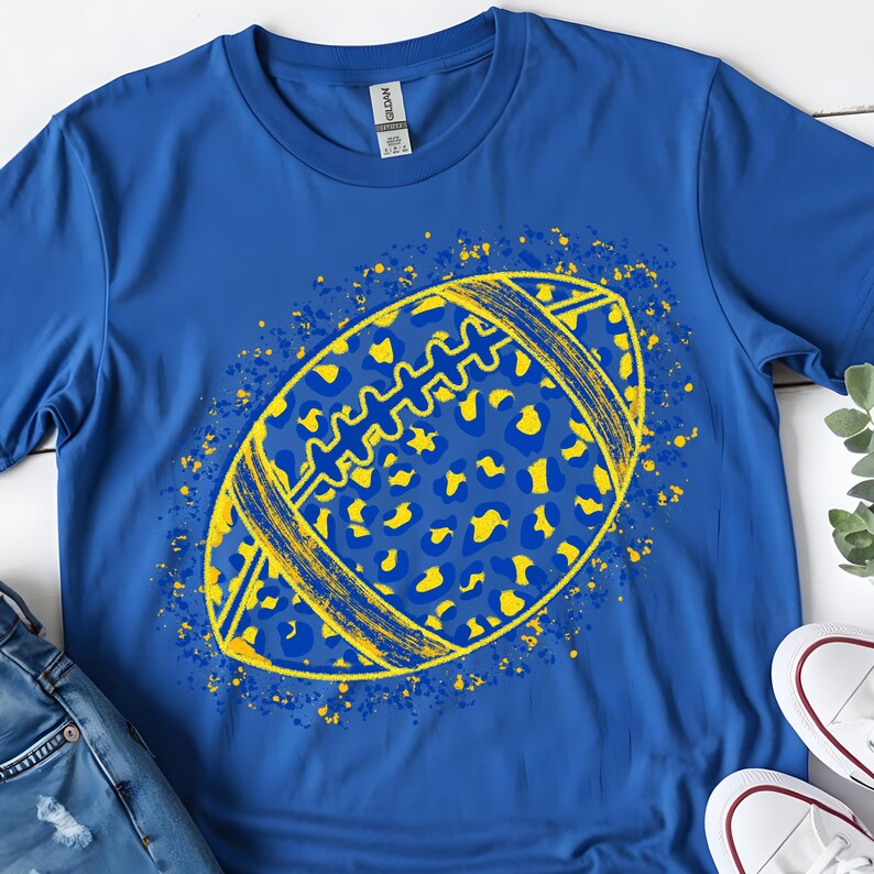May include: A royal blue short-sleeve t-shirt featuring a central graphic of an American football. The football has a yellow and blue leopard print pattern with yellow paint splatter accents surrounding the shape of the ball.