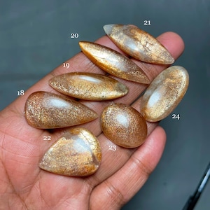 May include: Seven sunstone cabochons in various shapes and sizes, with a golden shimmer and a translucent appearance. The stones are numbered 18 through 24.