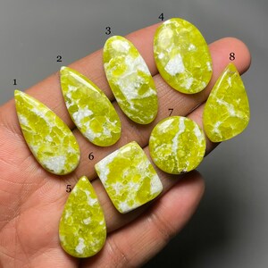 May include: Eight green and white cabochon gemstones with a mottled pattern. The gemstones are arranged in a row on a hand.