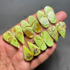 May include: Six pairs of green and yellow opal cabochons, each with a unique pattern and color variation. The cabochons are shaped like teardrops and are arranged in a row on a hand.