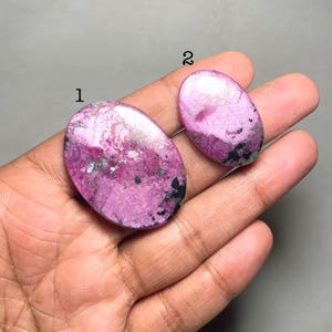 Może przedstawiać: Dwa owalne kamienie szlachetne o wypolerowanym wykończeniu. Większy kamień jest w żywym różowym kolorze z czarnymi i białymi wtrąceniami. Mniejszy kamień ma podobne zabarwienie. Kamienie szlachetne są prezentowane na dłoni na szarym tle.