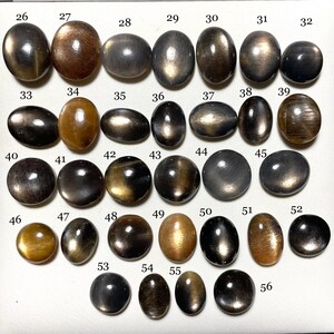 May include: A collection of 56 oval and round cabochon gemstones, with a variety of colours, including black, brown, and gold. The stones are arranged in rows and numbered from 26 to 56.