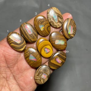 Könnte beinhalten: Eine Sammlung ovaler Edelsteine mit einem braunen und beigen Holzmaserungsmuster. Jeder Stein hat einen lebendigen, schillernden Opalmittelpunkt, der ein Farbspektrum zeigt. Die Edelsteine werden vor einem dunklen Hintergrund präsentiert.