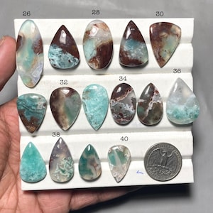 May include: A collection of polished gemstones in various shapes, including oval, teardrop, and triangular forms. The stones display a mix of colors, such as teal, brown, and white, with unique patterns and textures. A quarter coin is included for size reference.