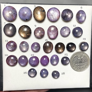 May include: A collection of polished gemstones in various shades of purple, brown, and gray, displayed on a white surface. The gemstones vary in size and shape, with some being round and others oval. A US quarter coin is included for size comparison.