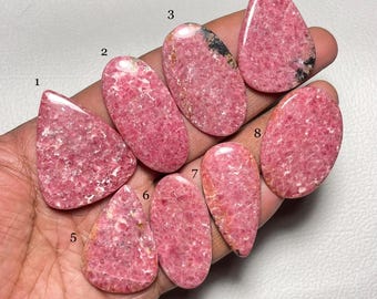Puede incluir: Ocho cabujones de rodocrosita rosa y blanca, cada uno con un patrón y una forma únicos, se muestran en una mano. Los cabujones están numerados del 1 al 8.