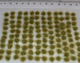 Op de afbeelding: Een vel met 100 groene en bruine plukjes statisch gras, gerangschikt in een rasterpatroon op een wit oppervlak. De plukjes zijn ongeveer 1,25 cm in diameter.