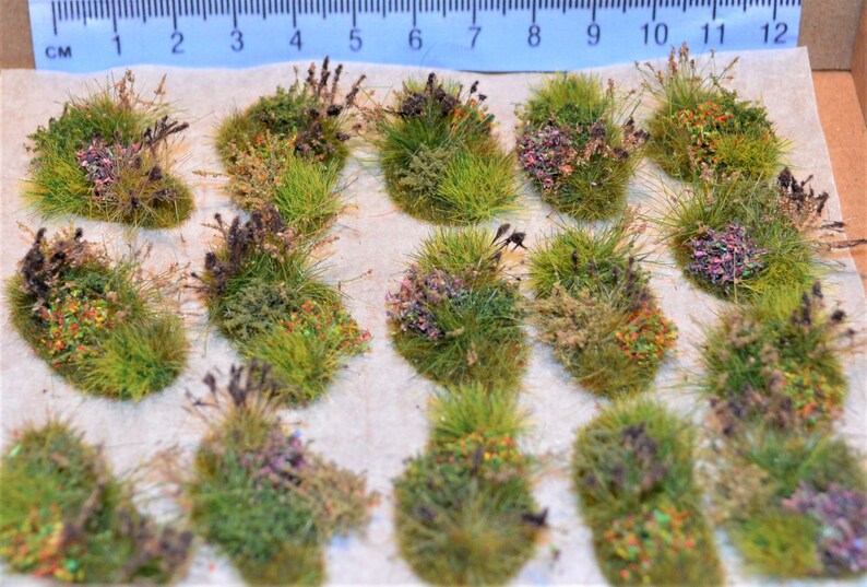 Puede incluir: Un conjunto de 16 mechones de hierba en miniatura de color verde, marr&oacute;n y morado con acentos coloridos. Los mechones est&aacute;n dispuestos en un patr&oacute;n de cuadr&iacute;cula sobre una superficie blanca.