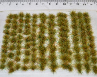 Op de afbeelding: Een vel met groene, bruine en gele statische gras plukjes, gerangschikt in rijen op een witte achtergrond. De plukjes zijn ongeveer 1,2 cm lang.
