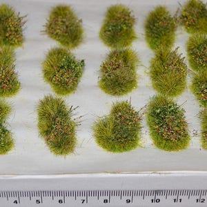 Puede incluir: Una colecci&oacute;n de 15 peque&ntilde;os mechones de hierba artificial verde y marr&oacute;n con flores rojas y amarillas. Los mechones est&aacute;n dispuestos en una cuadr&iacute;cula sobre una superficie blanca.