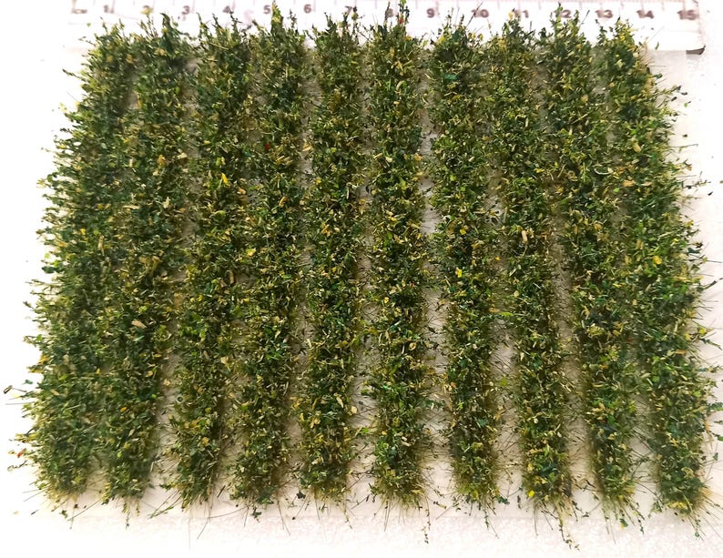 May include: A sheet of green and yellow model railroad grass tufts, arranged in rows on a white surface. The tufts are approximately 1 inch in length.