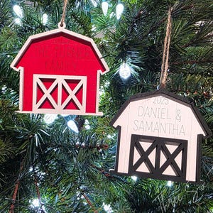 May include: Two wooden barn-shaped Christmas ornaments hanging on a tree. One is red with white accents and the text "THE TURNER FAMILY". The other is white with black accents and the text "2025 DANIEL & SAMANTHA".