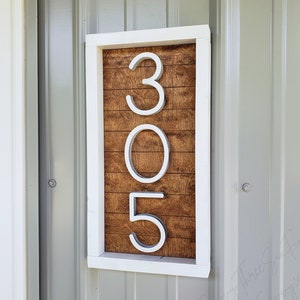 May include: A rectangular wooden address sign with the numbers "305" in white, set against a brown wood-grain background. The sign is framed in white and mounted on a gray metal surface.