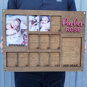 May include: Wooden baby milestone photo frame with spaces for photos from newborn to one year. The frame includes the name "Parker Rose" in pink letters. The frame has spaces for photos labeled "Newborn", "Three Months", "Four Months", "Five Months", "Six Months", "Seven Months", "Eight Months", "Nine Months", "Ten Months", "Eleven Months", and "First Birthday".