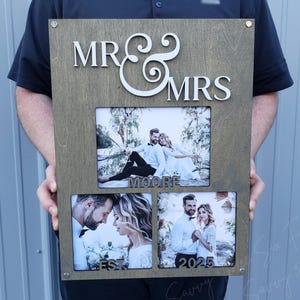 May include: A wooden photo frame with the words "MR & MRS" and "MOORE" in white lettering. The frame has three openings for photos, with one photo showing a couple sitting on a tree branch, another photo showing a couple looking at each other, and the third photo showing a couple standing together. The frame also has the text "EST 2025" in white lettering.