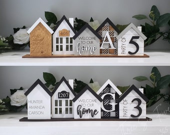 May include: A set of five wooden house-shaped ornaments with black and white details. The first house has the names James, Sarah, Kendra, Austin, and Bella. The second house has the number 406. The third house says "Welcome to our home". The fourth house has the letter A. The fifth house says "Party of 5". The second set of houses has the names Hunter, Amanda, and Carson. The second house has the number 1207. The third house says "Welcome to our home". The fourth house has the letter G. The fifth house says "Party of 3".