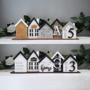 May include: A set of five wooden house-shaped ornaments with black and white details. The first house has the names James, Sarah, Kendra, Austin, and Bella. The second house has the number 406. The third house says "Welcome to our home". The fourth house has the letter A. The fifth house says "Party of 5". The second set of houses has the names Hunter, Amanda, and Carson. The second house has the number 1207. The third house says "Welcome to our home". The fourth house has the letter G. The fifth house says "Party of 3".