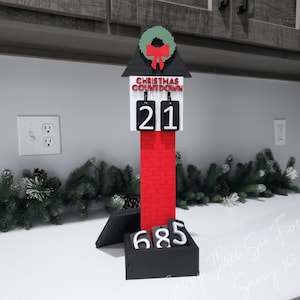 May include: A black and red Christmas countdown calendar with a white house-shaped top, a wreath, and a red bow. The calendar displays the numbers 21 and 685.