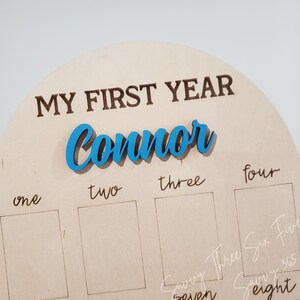 May include: A wooden plaque with the words "My First Year" and the name "Connor" in blue lettering. The plaque has eight empty spaces for photos, labeled with the numbers one through eight.