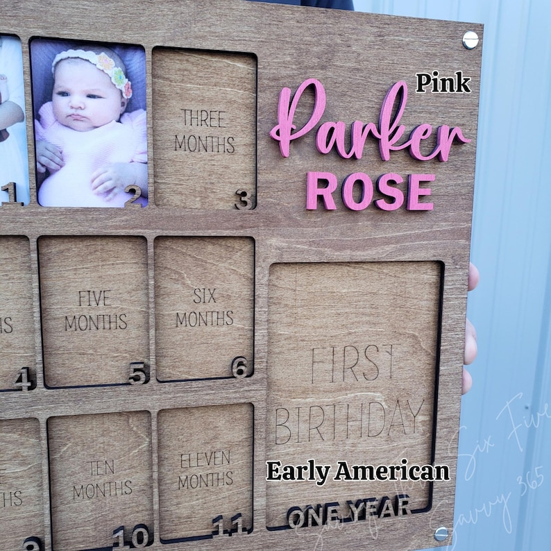 May include: Wooden baby milestone photo frame with spaces for photos and months. Features the name "Parker Rose" in pink, with text for "First Birthday" and "Early American". Includes spaces for photos from 1 to 11 months.