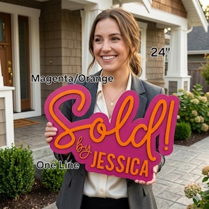 May include: A magenta and orange "Sold by Jessica" sign, 24 inches in length, held by a person. The sign features the word "Sold!" in orange script over the magenta base, with "by Jessica" in a smaller font. The sign is displayed in front of a house.