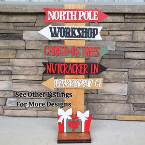 May include: Wooden Christmas-themed directional sign with arrows pointing to "North Pole", "Workshop", "Christmas Trees", "Nutcracker IN", and "Gingerbread CT". The sign includes gift box cutouts at the base and text that reads "See Other Listings For More Designs".